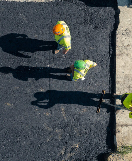 Three workers in safety gear spread asphalt, casting long shadows.