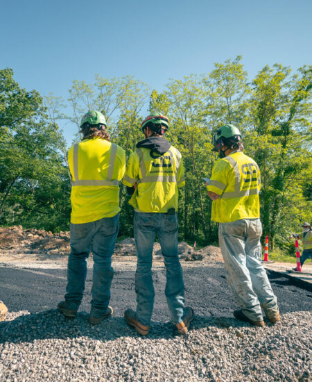 Workers in yellow vests and helmets on gravel at outdoor site.