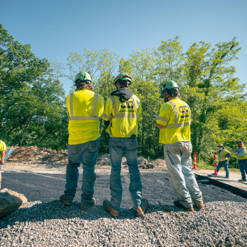 Workers in yellow vests and helmets on gravel at outdoor site.