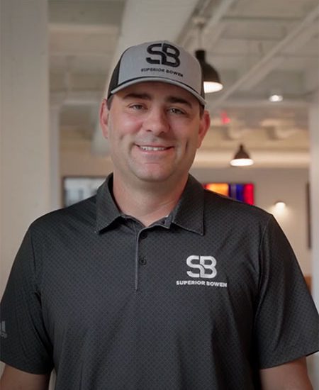 Man in black Superior Bowen polo and cap smiles indoors.