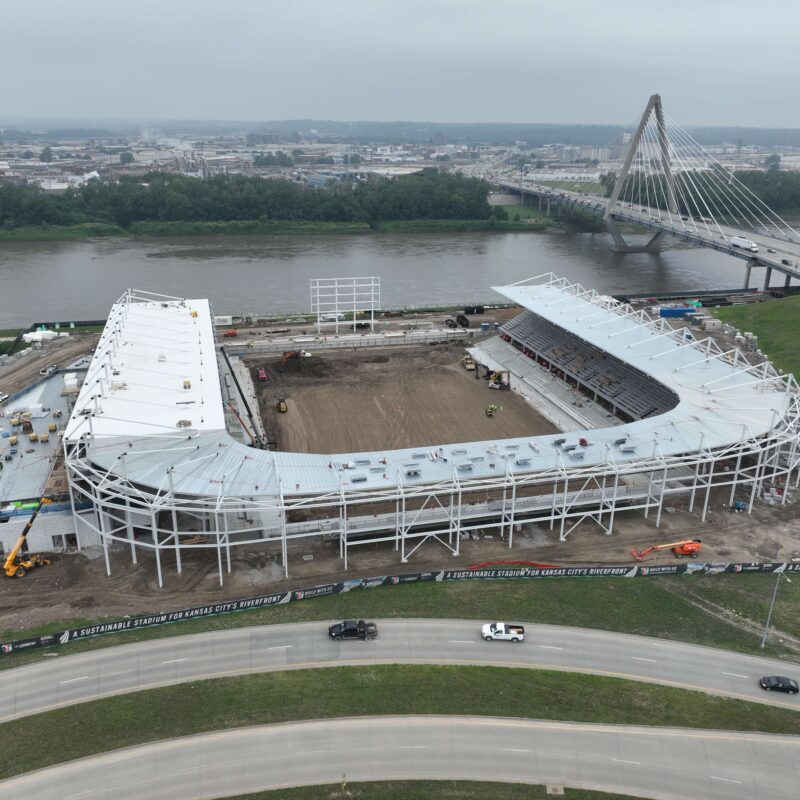 Stadium construction by river, bridge and city in background.