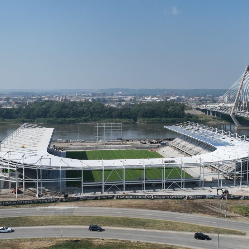 Partially built stadium by river and bridge, cars in front.