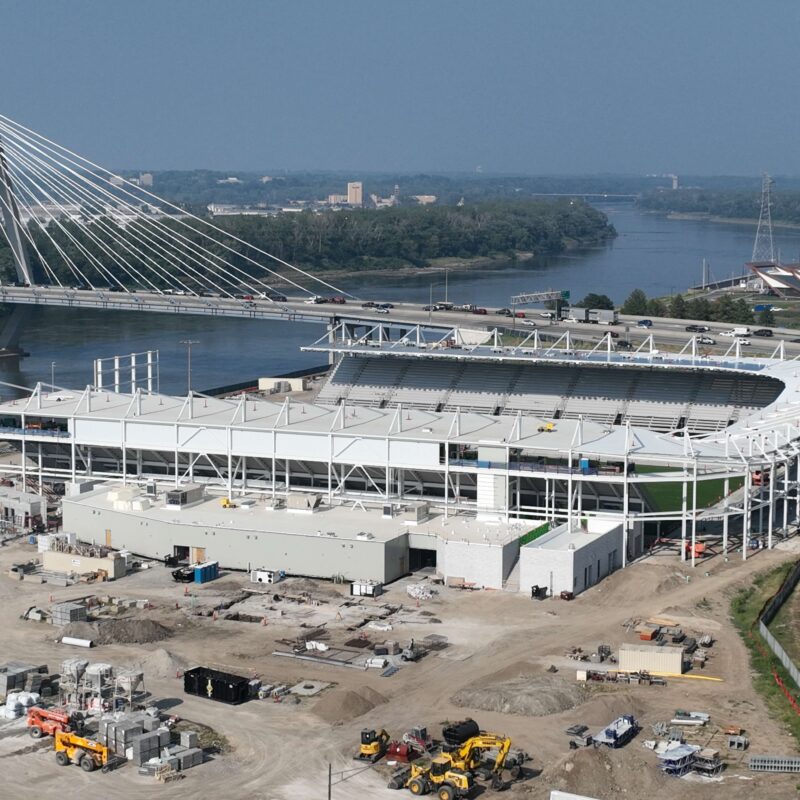 Stadium under construction by river, with bridge and vehicles nearby.