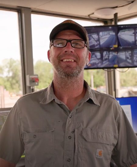 Man in glasses and cap stands in control room with monitors.