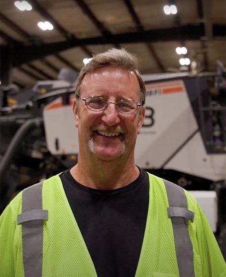 Smiling man in neon vest and glasses stands by industrial equipment.