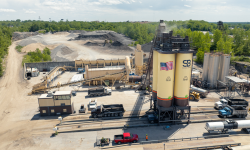 Aerial view of asphalt plant with silos, trucks, and trees.