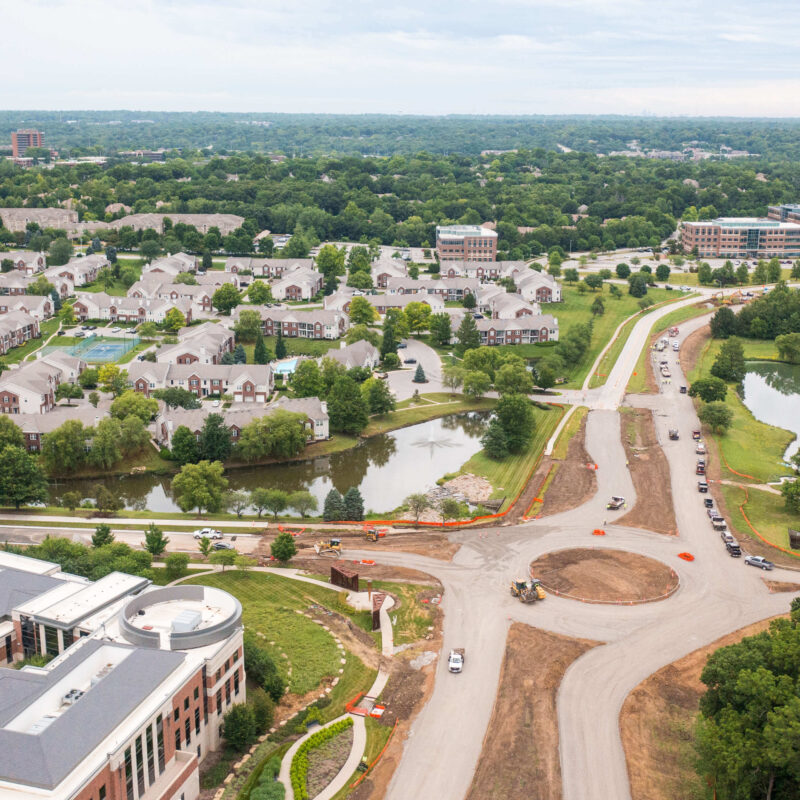 Suburban aerial view: homes, pond, trees, roadwork with vehicles.