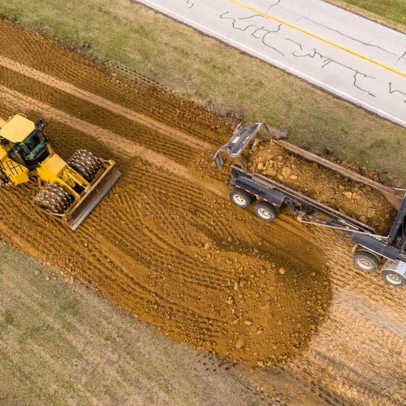 Bulldozer spreads dirt near dump truck at roadside construction.