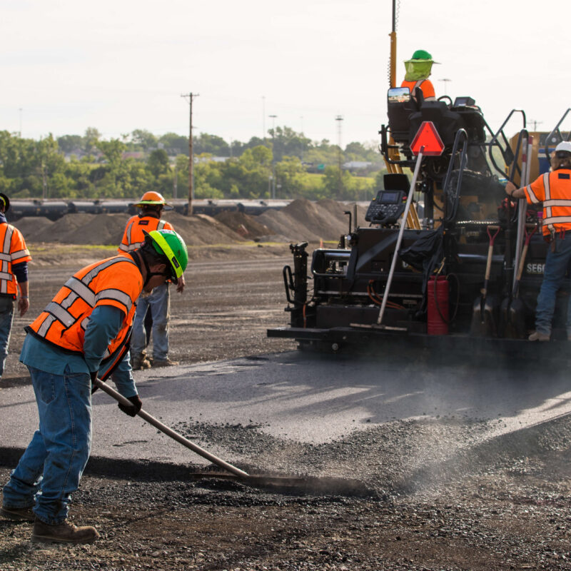 Workers in safety gear smooth asphalt beside paving machine.