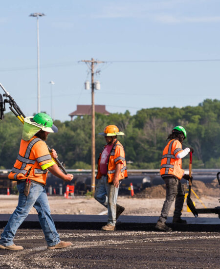Workers in safety gear pave road, one carries equipment.