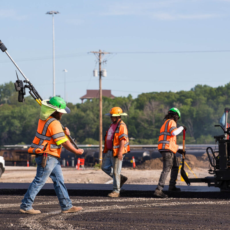 Workers in safety gear pave road, one carries equipment.