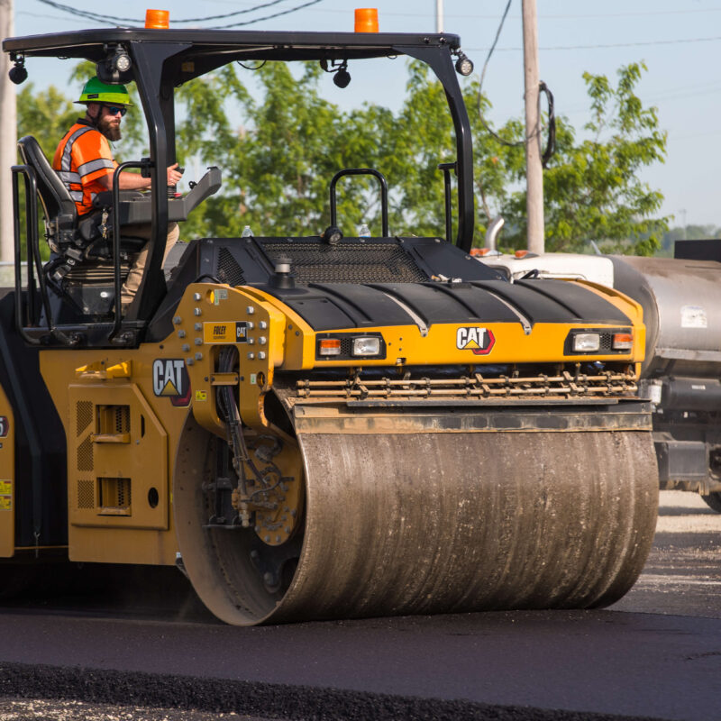 Worker drives yellow CAT roller smoothing fresh pavement.