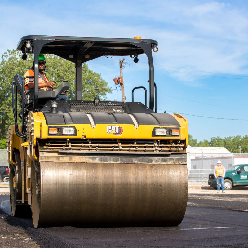 Worker uses yellow CAT steamroller to flatten road asphalt.
