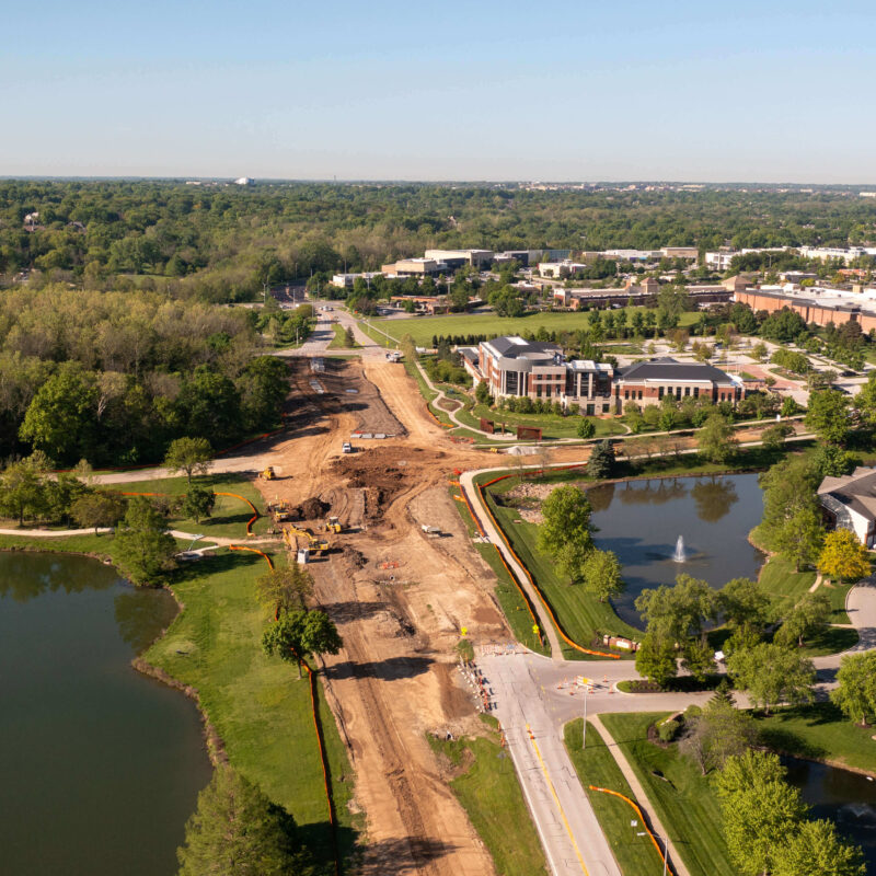 Aerial view: construction site, heavy machinery, pond, homes, trees.