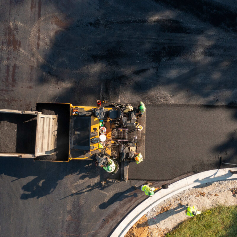 Workers operate machines to lay asphalt; truck supplies material.