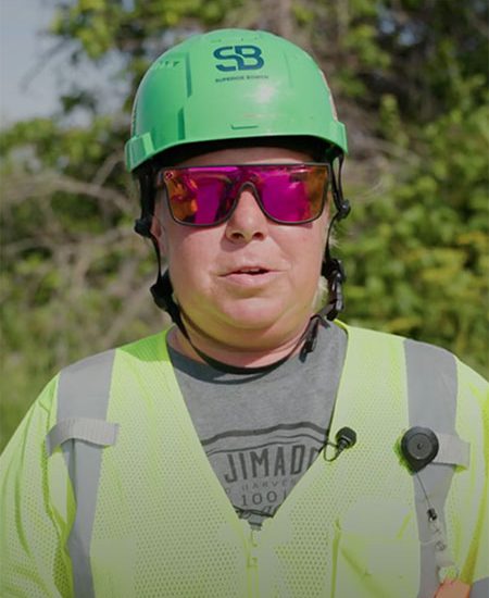 Person in green safety helmet, vest, and pink sunglasses outdoors.