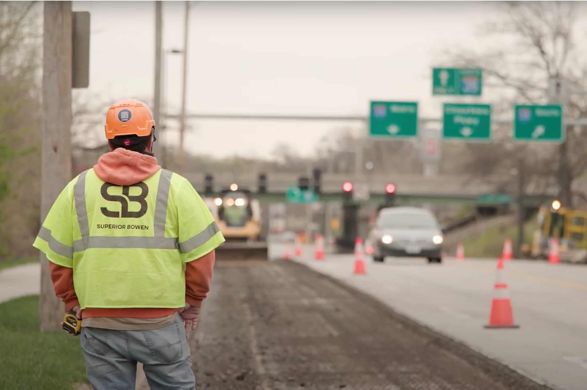 Construction worker in vest by roadwork, cones and signs behind.