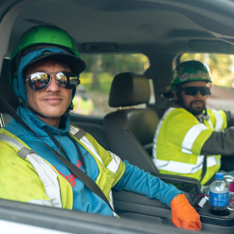 Two workers in safety gear sit in a vehicle.
