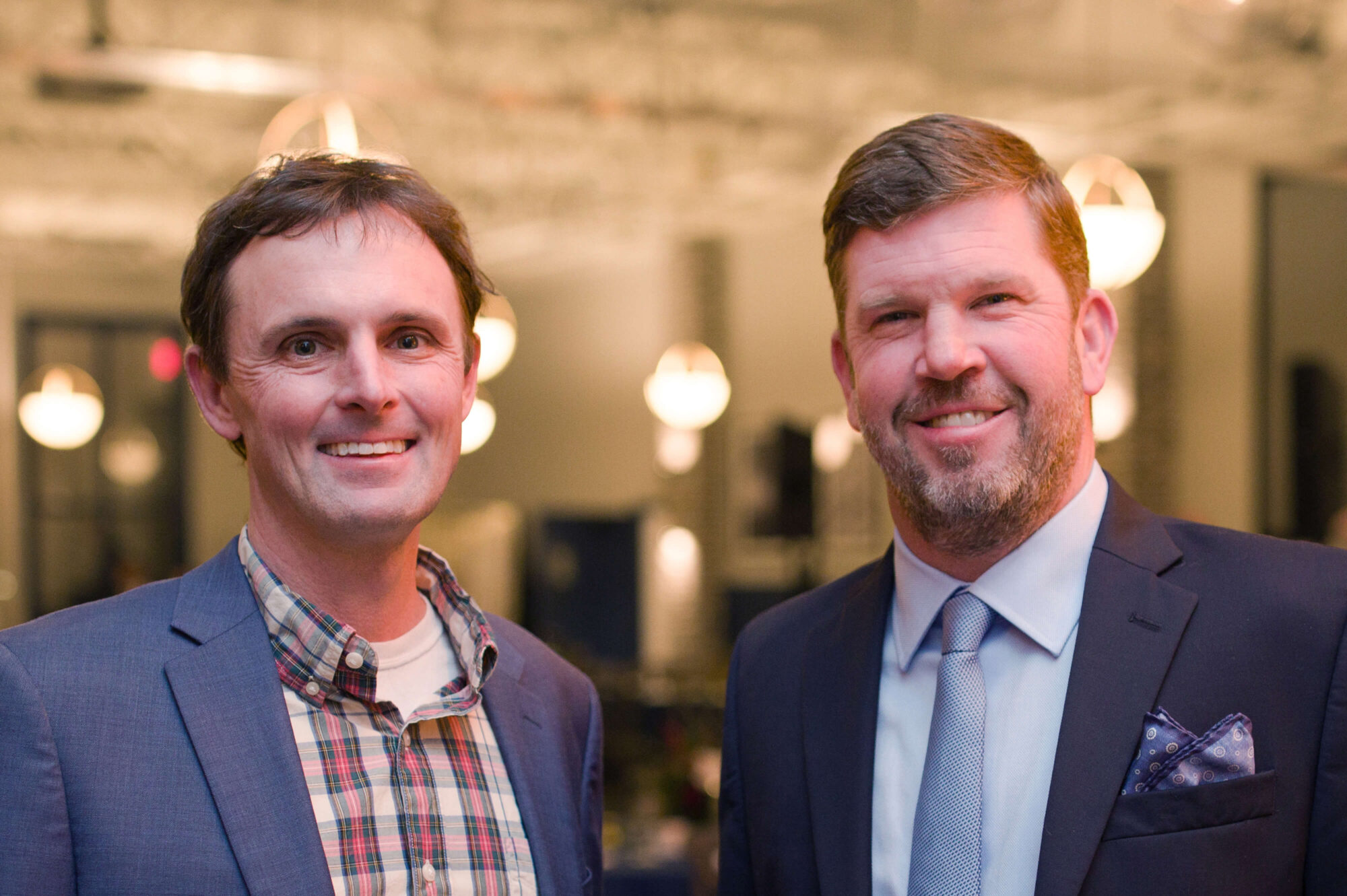 Two smiling men indoors, one in plaid and blazer, one in suit.
