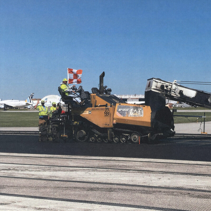 Workers use a paving machine on an airport runway.