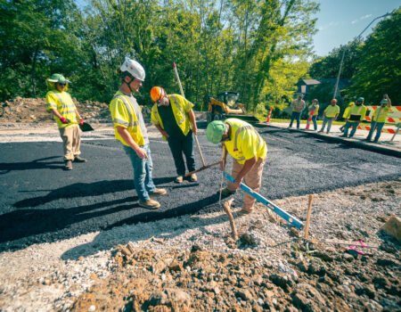 Workers in safety gear pave road, measure, surrounded by machines.
