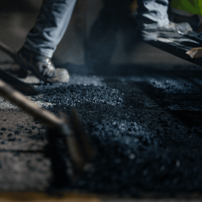 Workers' feet and tools spreading fresh, dark asphalt on road.