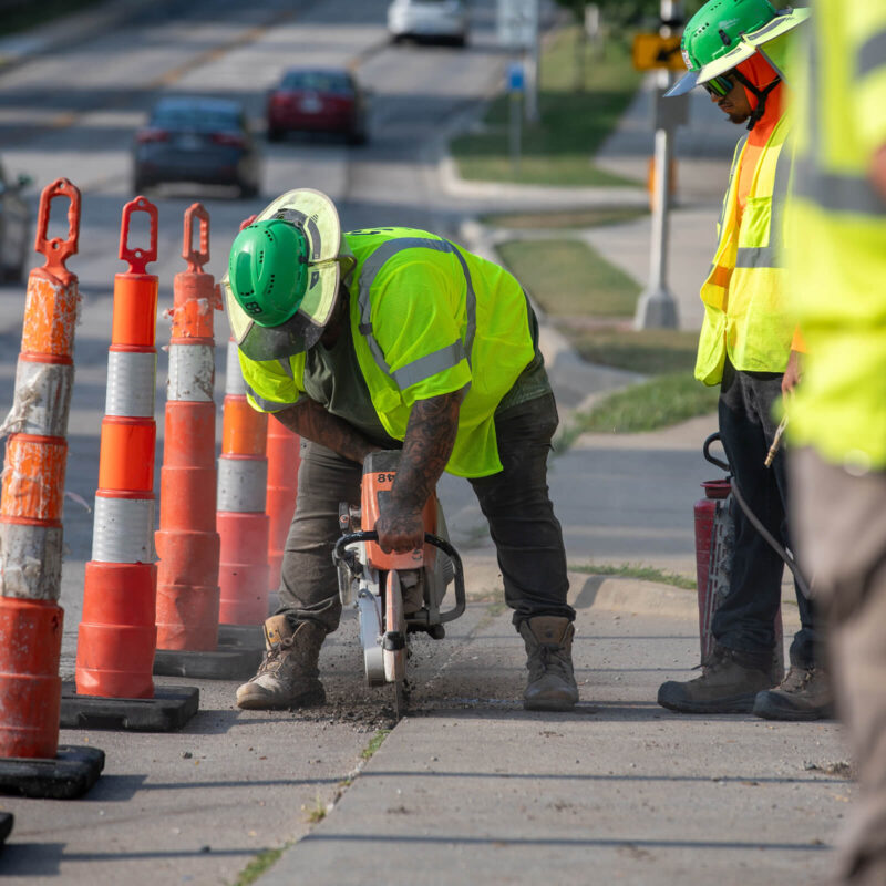 Worker in safety gear cuts sidewalk with concrete saw.