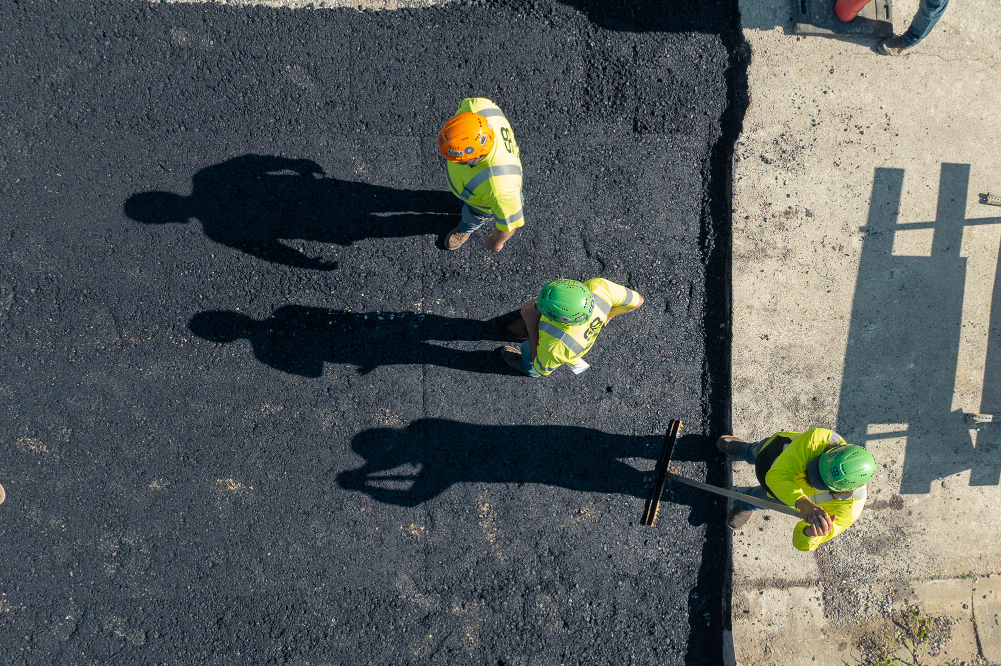 Workers in helmets spread asphalt; shadows on pavement.