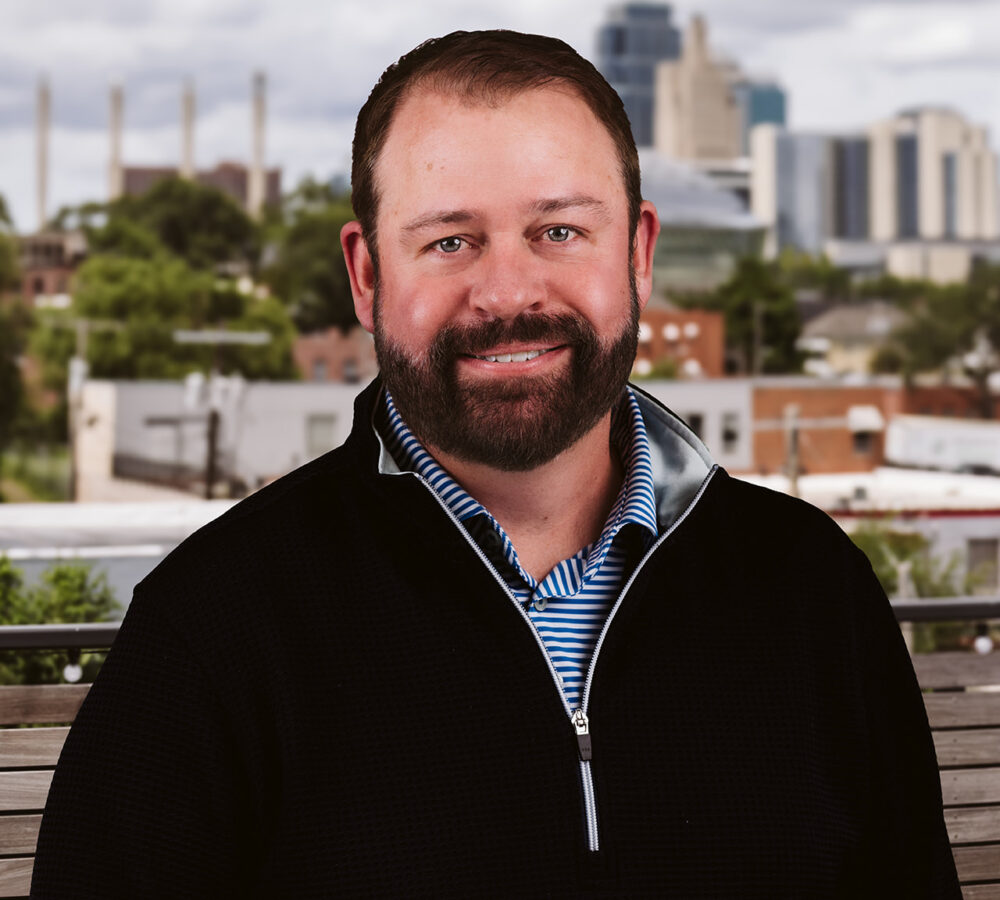 Bearded man in dark sweater outside with urban skyline.