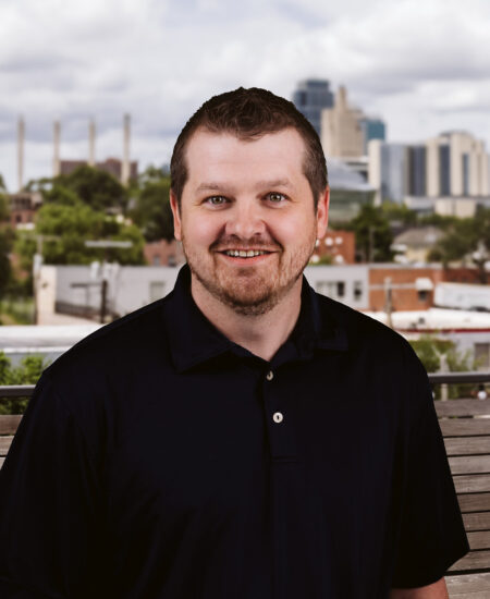 Bearded man in black polo shirt outdoors, cityscape behind.
