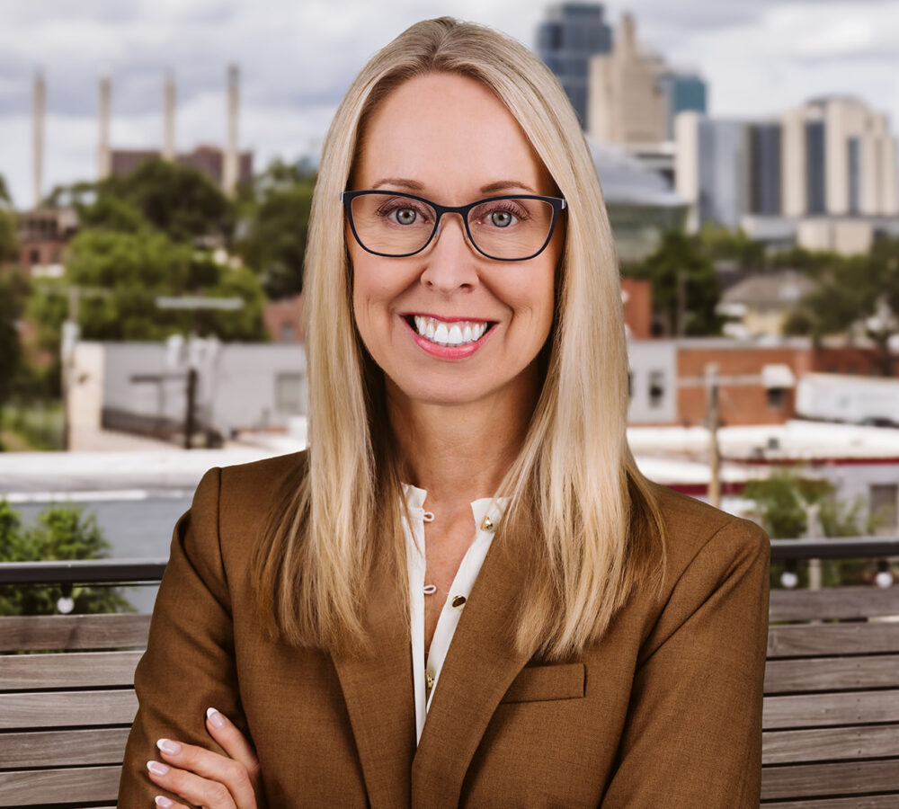 Blonde woman in glasses and brown suit stands outside.