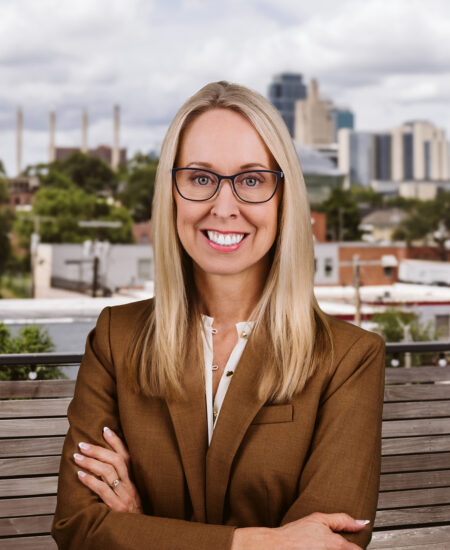 Blonde woman in glasses and brown suit stands outside.
