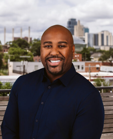 Smiling man on outdoor bench, cityscape and clouds behind.