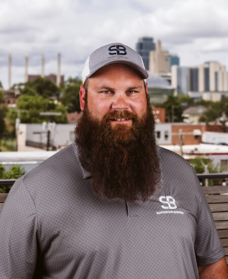 Bearded man in gray Superior Bowen polo and cap, city backdrop.
