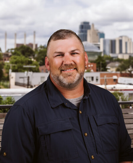 Smiling bearded man outdoors, city skyline behind him.