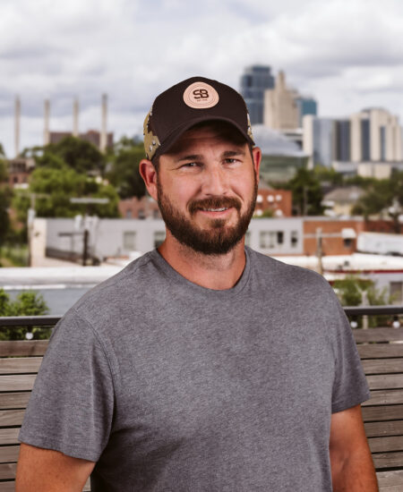 Bearded man in gray t-shirt and cap outside, city behind.