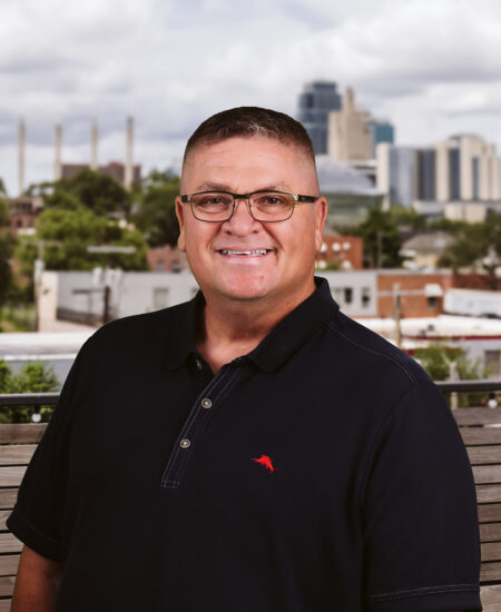 Man in glasses and dark polo shirt outdoors, city behind.