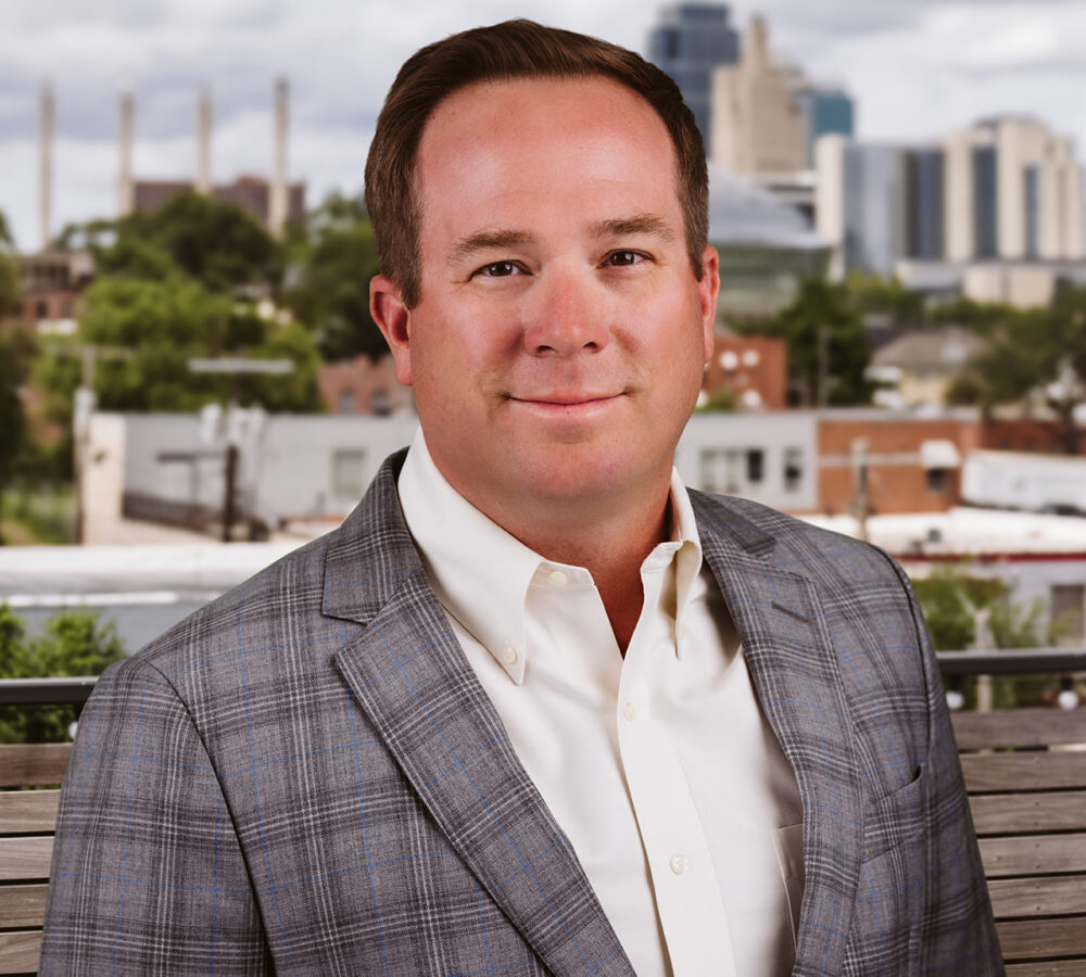 Man in plaid suit stands outside with city skyline behind.
