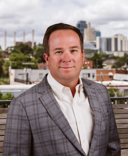Man in plaid suit stands outside with city skyline behind.