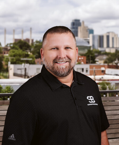 Bearded man in black polo shirt outdoors, city behind.