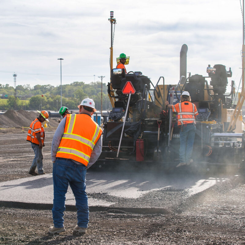 Workers in vests lay asphalt with machinery on a cloudy day.