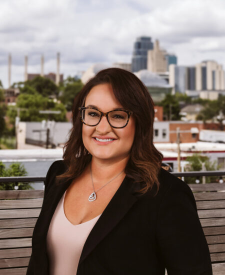 Smiling woman with glasses and brown hair on a bench, city behind.