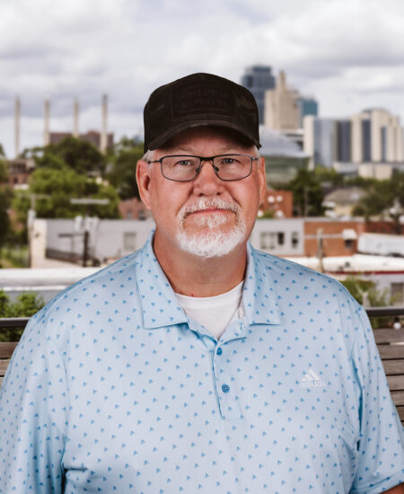 Man in glasses and cap outdoors, cityscape behind him.