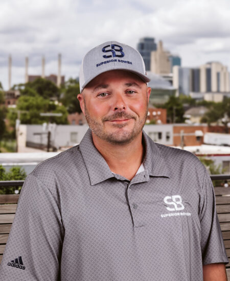 Man in gray Superior Bowen shirt and hat outdoors, city blurred.
