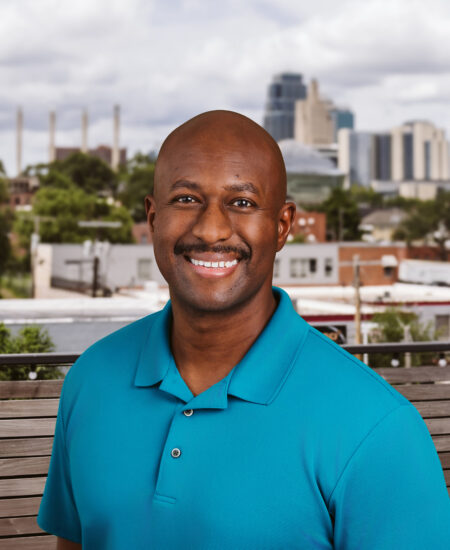 Smiling man in teal polo stands outdoors before city skyline.