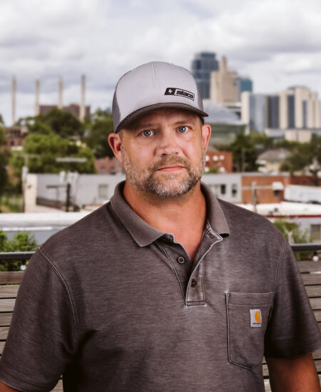 Bearded man in gray polo and cap outdoors, city behind.