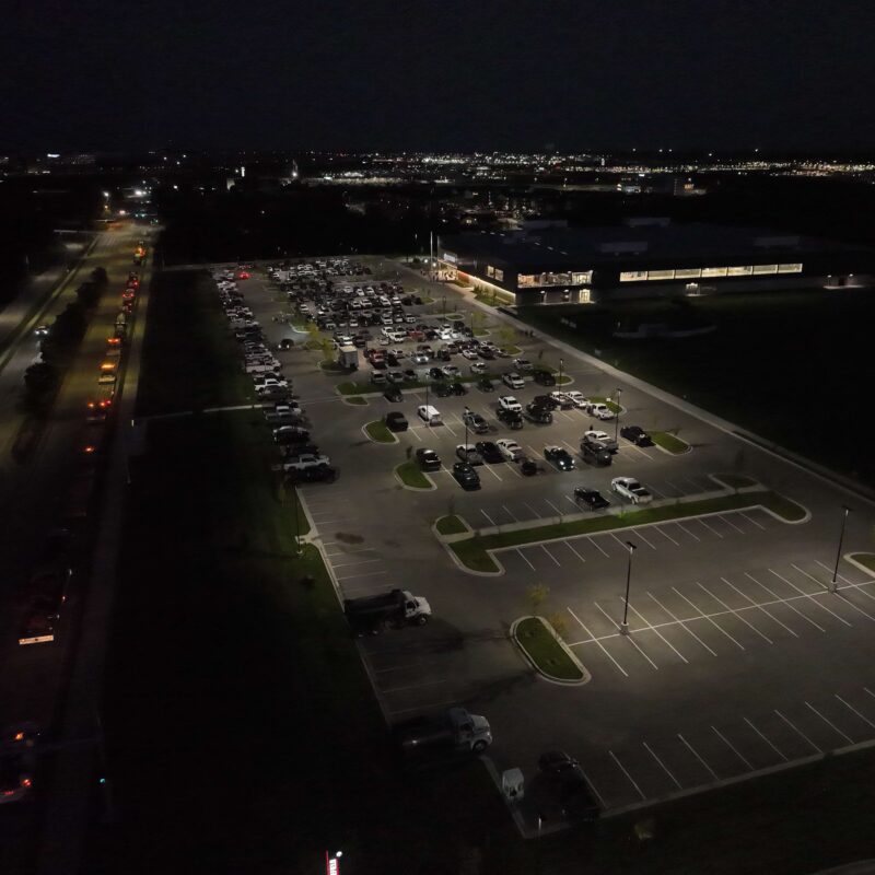 Aerial night view of lit parking lot, cars, nearby road.