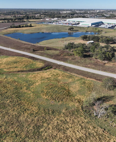 Winding road through grassy fields, pond, trees, and distant factories.