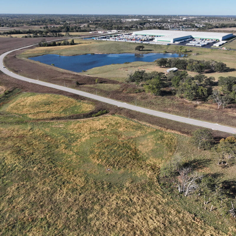Winding road through grassy fields, pond, trees, and distant factories.