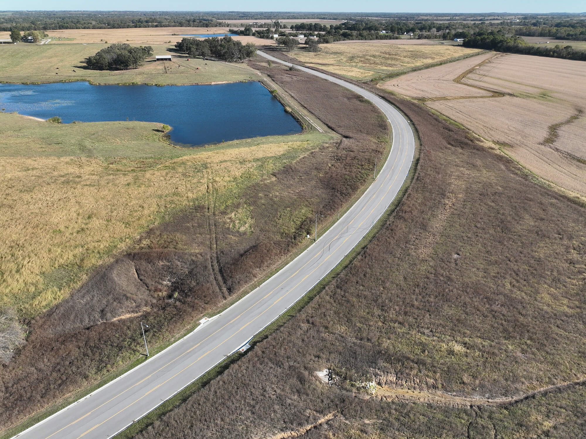 Winding road through grassy fields, pond, trees, and distant factories.